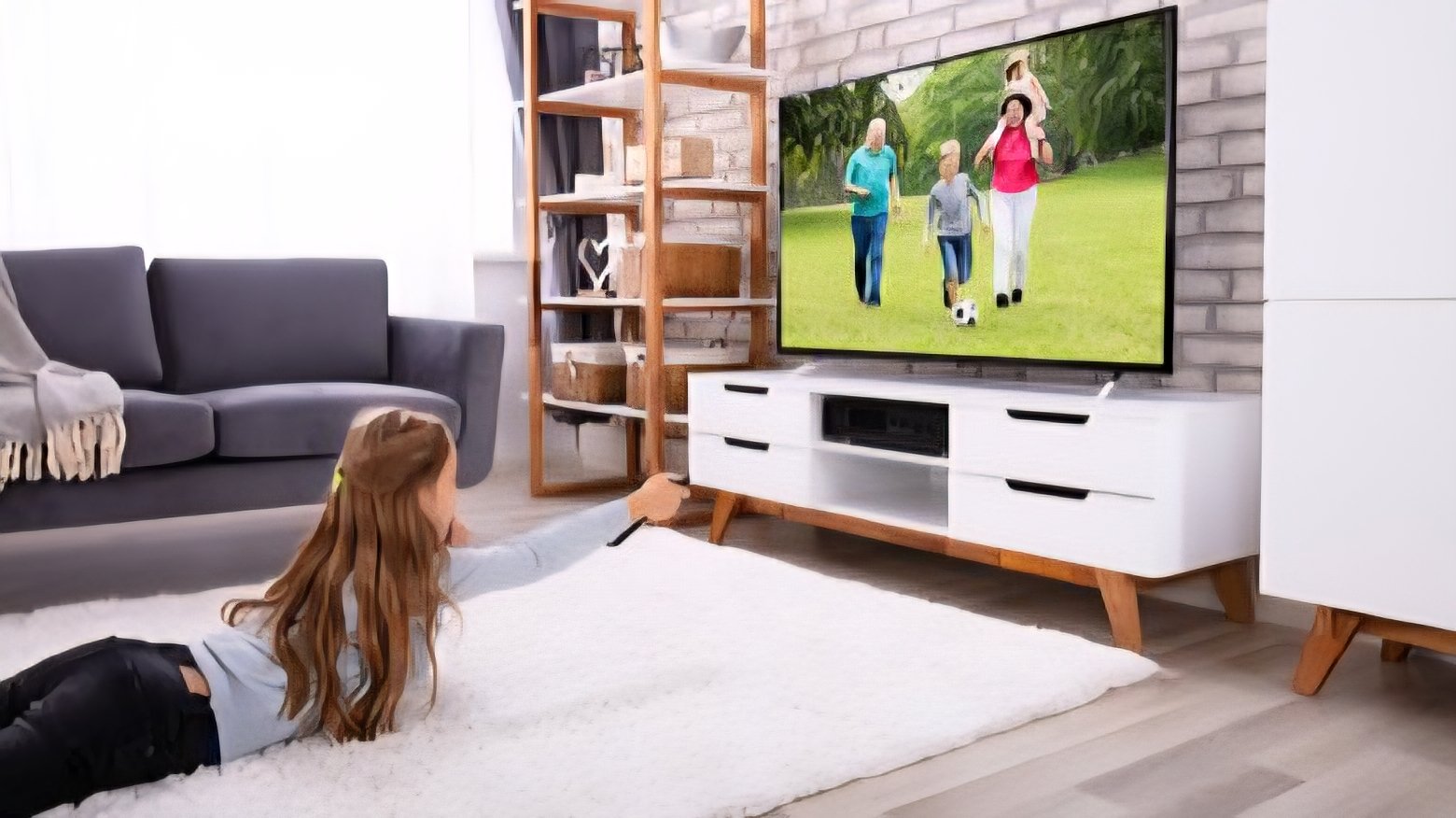 A young child lying on a white rug in a living room, holding a remote control and watching a television screen while a glass cabinet stands nearby.