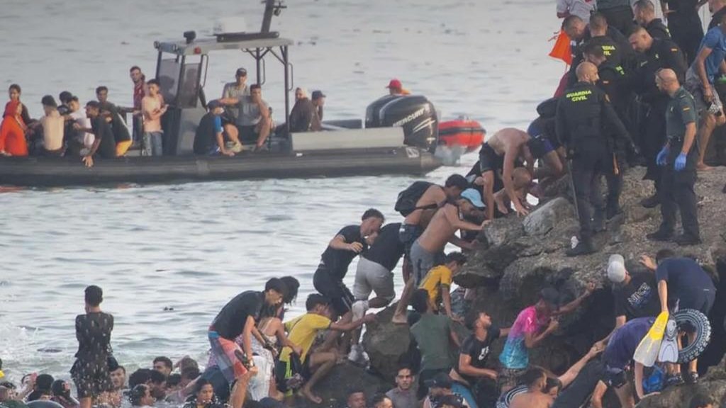 Migrants swimming near maritime border fence and gathering on beach in Ceuta enclave.