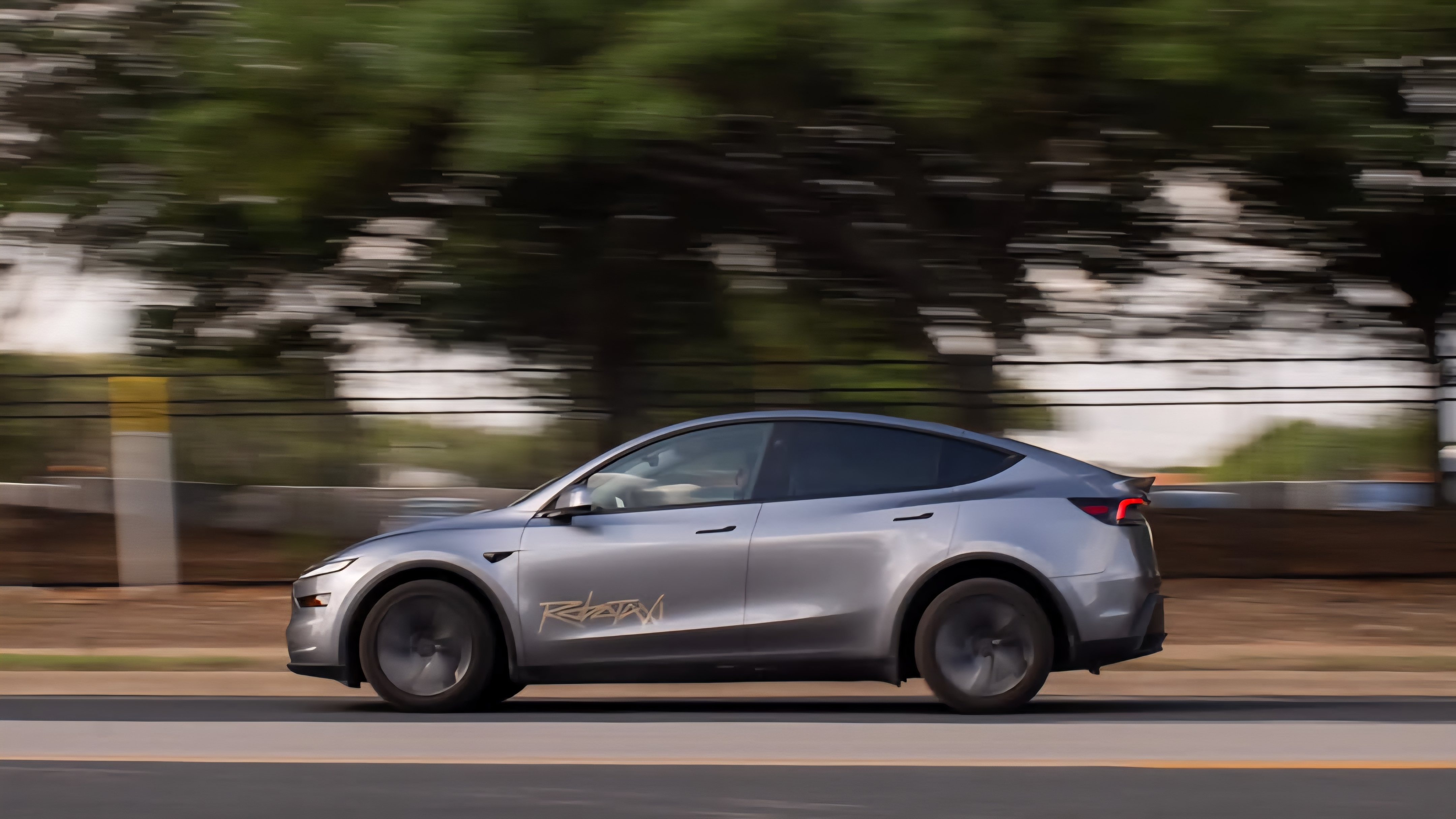 A silver Tesla Model Y robotaxi driving on an open asphalt road in Texas with green trees in the background.