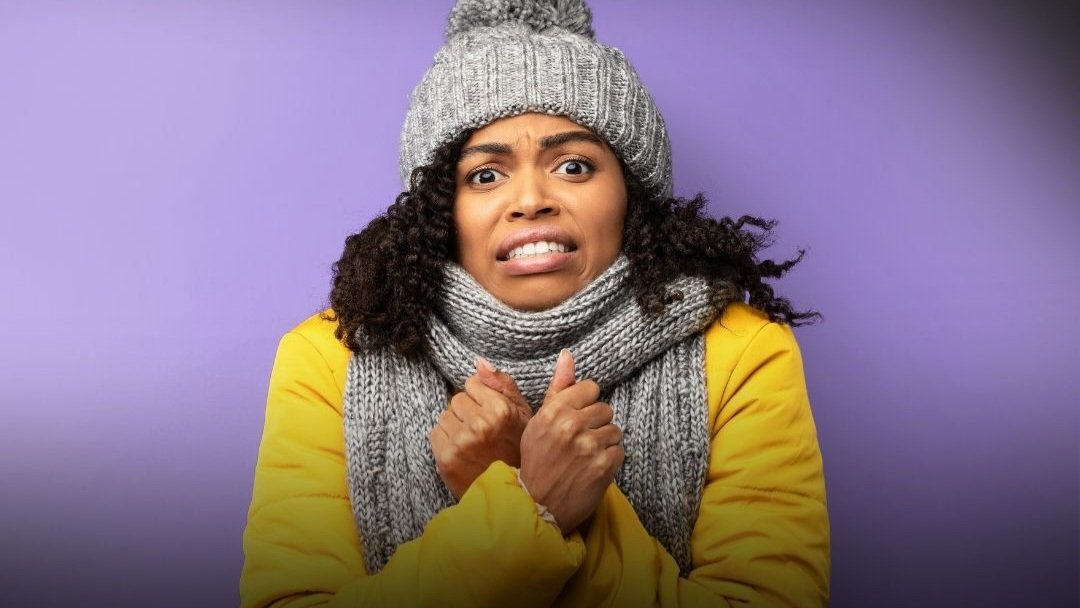 A young Black woman with curly hair, wearing a grey knit beanie, grey scarf, and yellow jacket, stands shivering against a solid purple background.