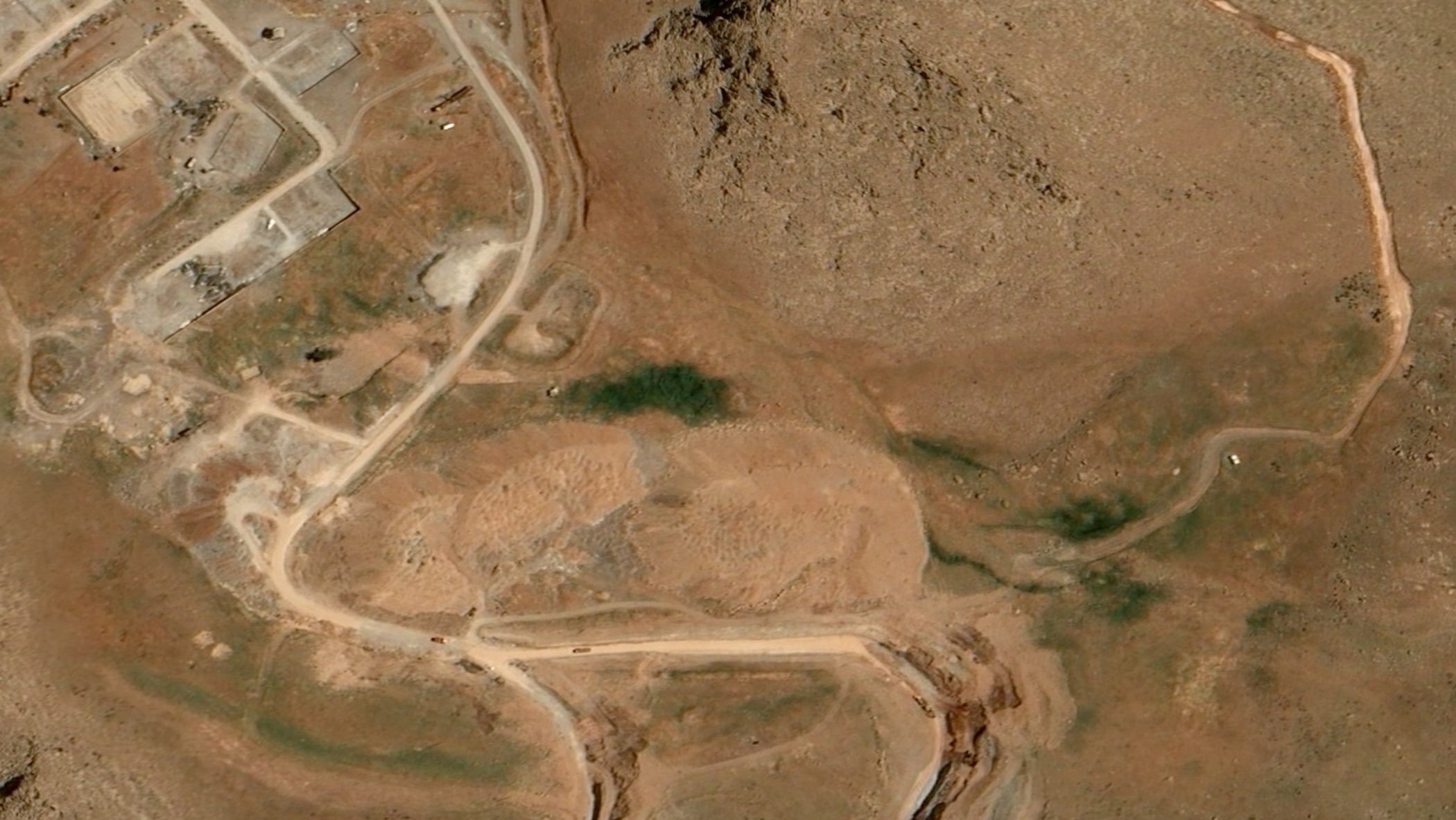 Satellite photograph showing an aerial view of arid, mountainous terrain with dirt access roads and circular earthwork structures in Iran.