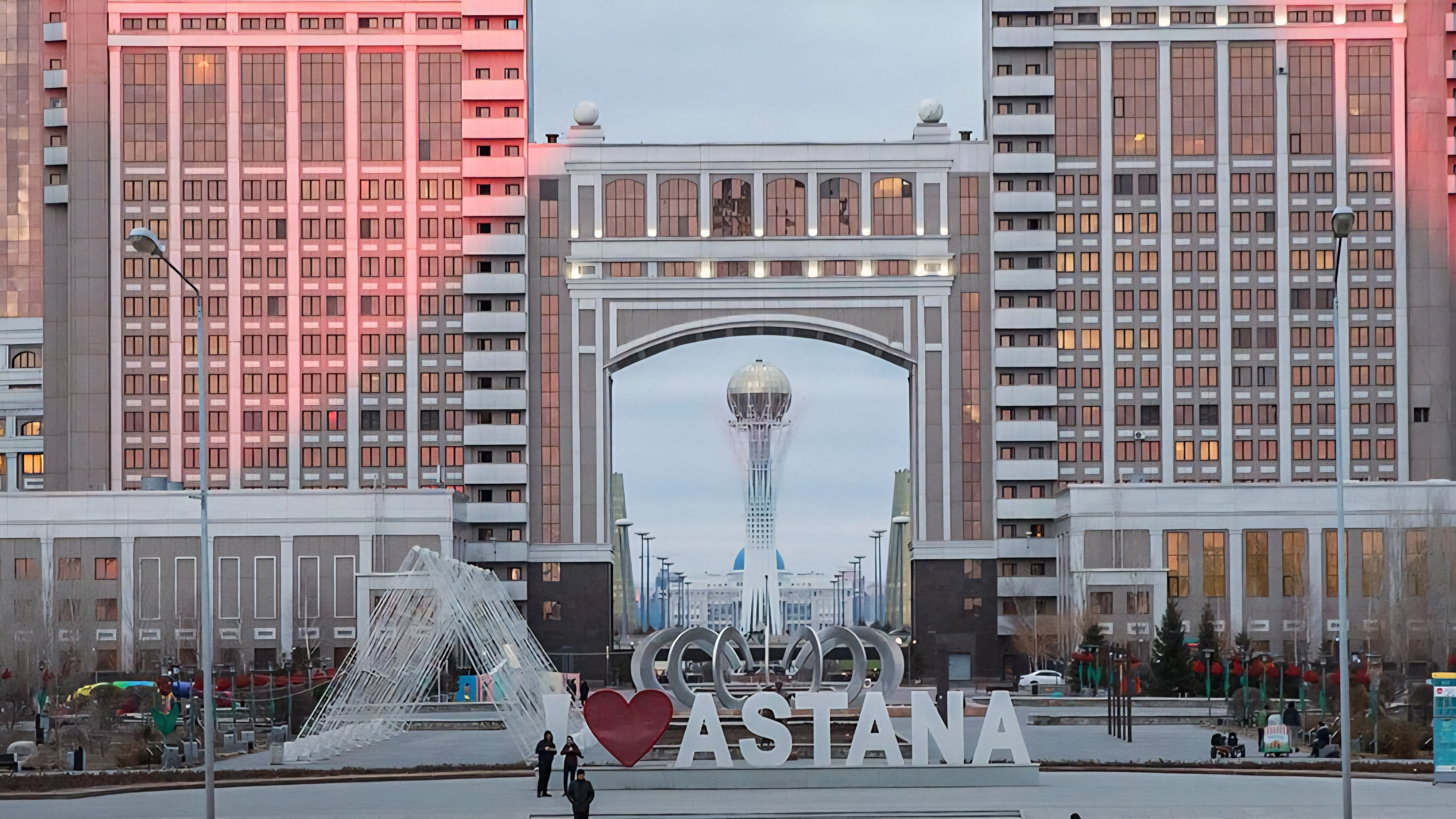 A wide view of the central square in Astana, Kazakhstan, showing the large monumental archway of the KazMunayGas headquarters building with the Bayterek Tower visible through the center opening.