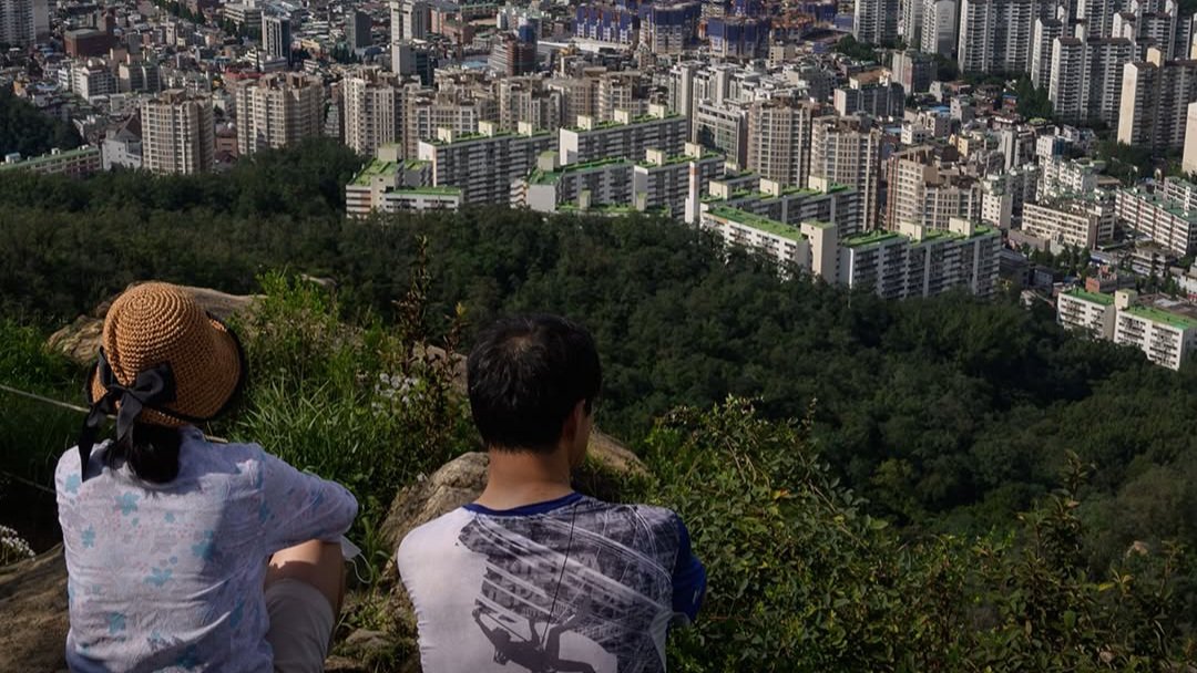 Two people sitting on a hill overlooking a dense urban cityscape with apartment blocks in South Korea.