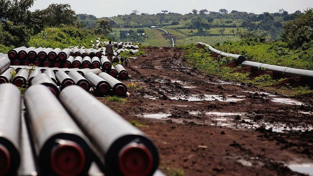 Steel pipes stacked beside a muddy pipeline construction corridor cutting through forested terrain.
