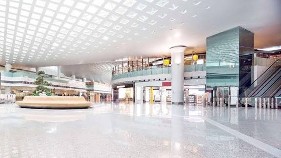 Architectural rendering of a modern, multi-level shopping mall interior with high ceilings, polished floors, and integrated indoor greenery.
