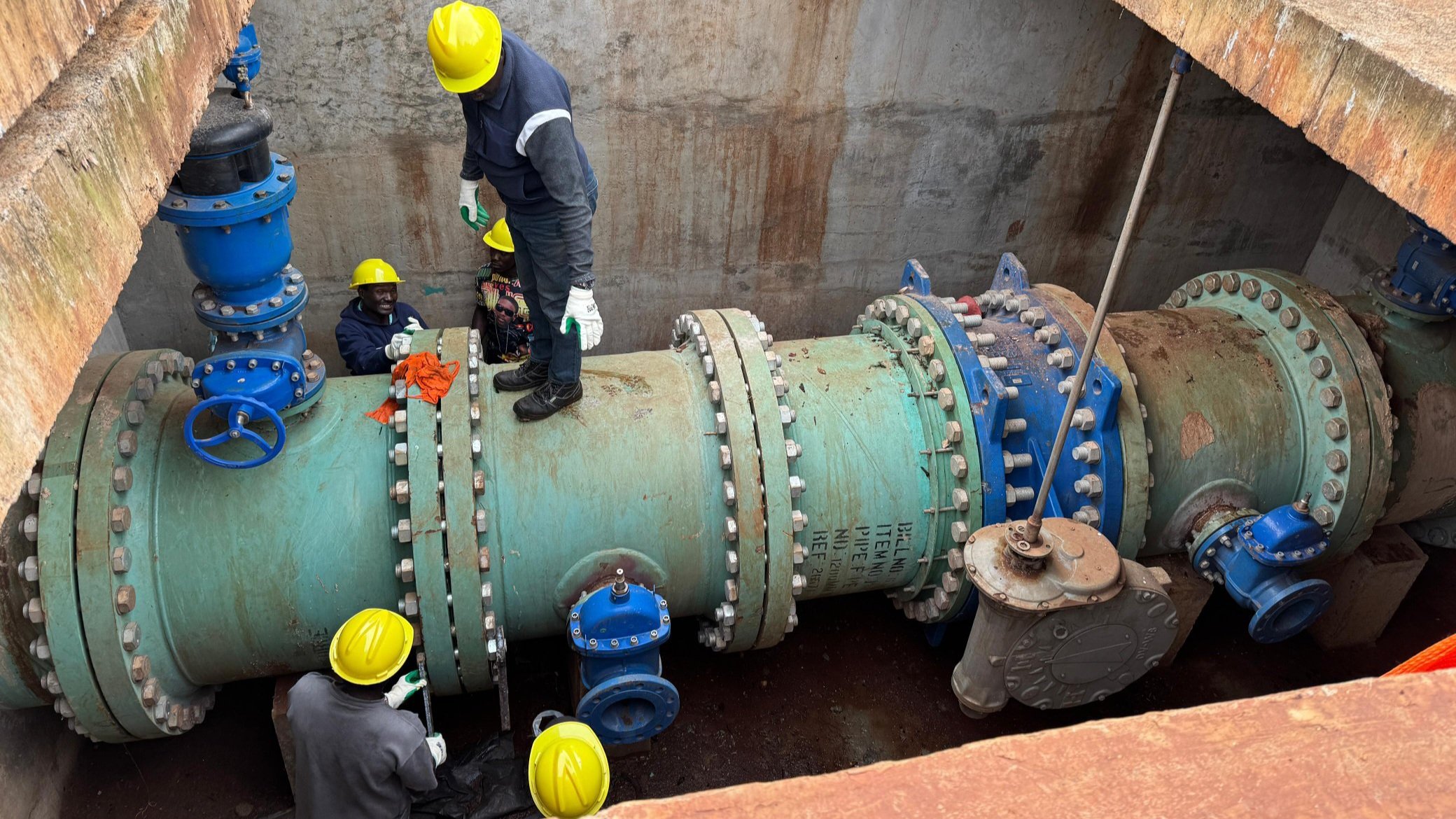 Construction workers and engineers wearing yellow helmets and high-visibility vests work on large green water pipes and blue valves inside a concrete chamber.