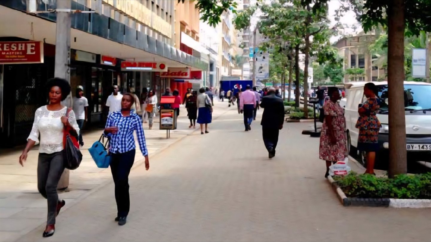 Pedestrians walking along a paved urban sidewalk lined with mature trees and store storefronts in Nairobi, Kenya.