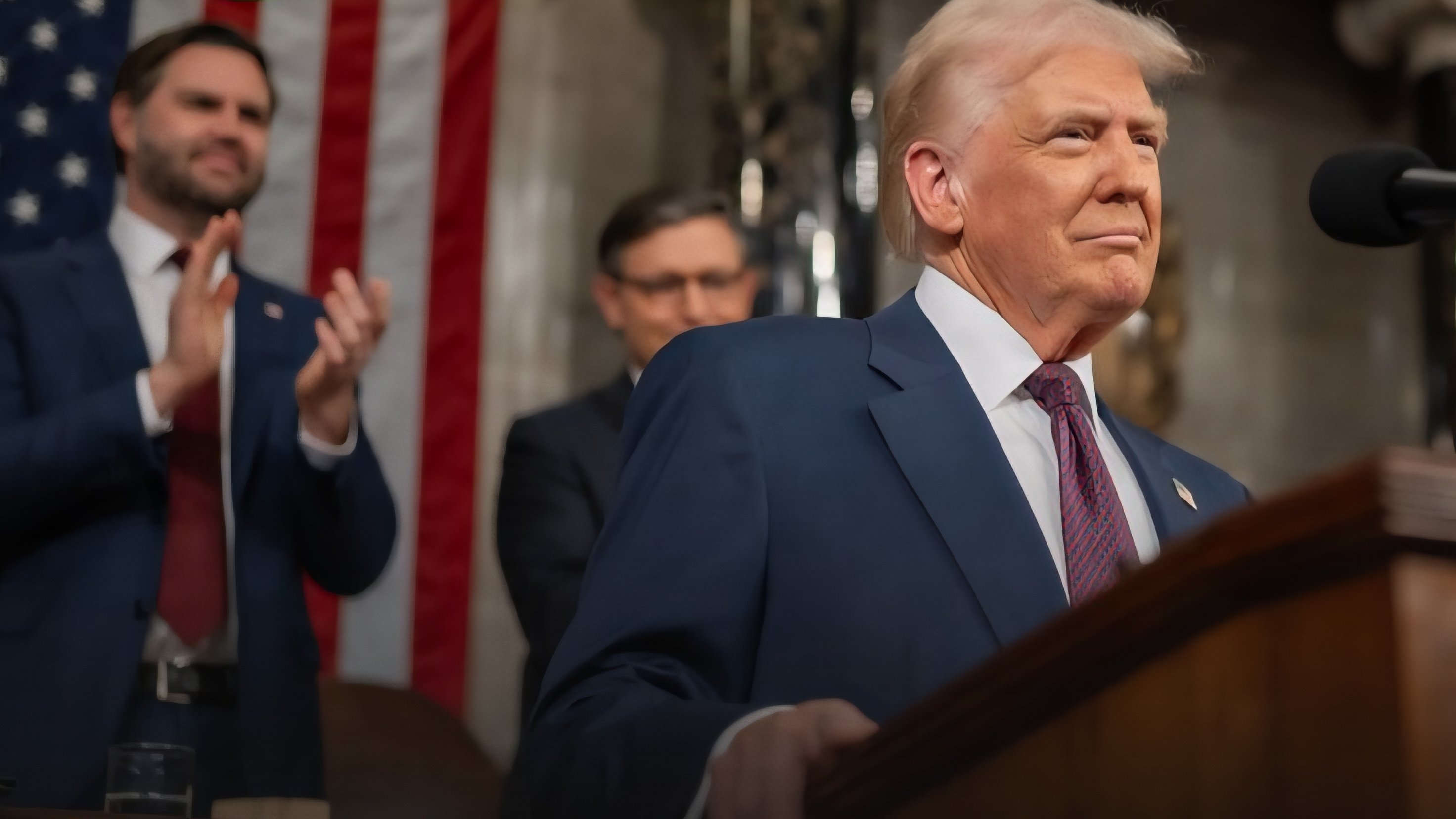 President Donald Trump speaking at a podium in front of an American flag with JD Vance and other officials applauding in the background.