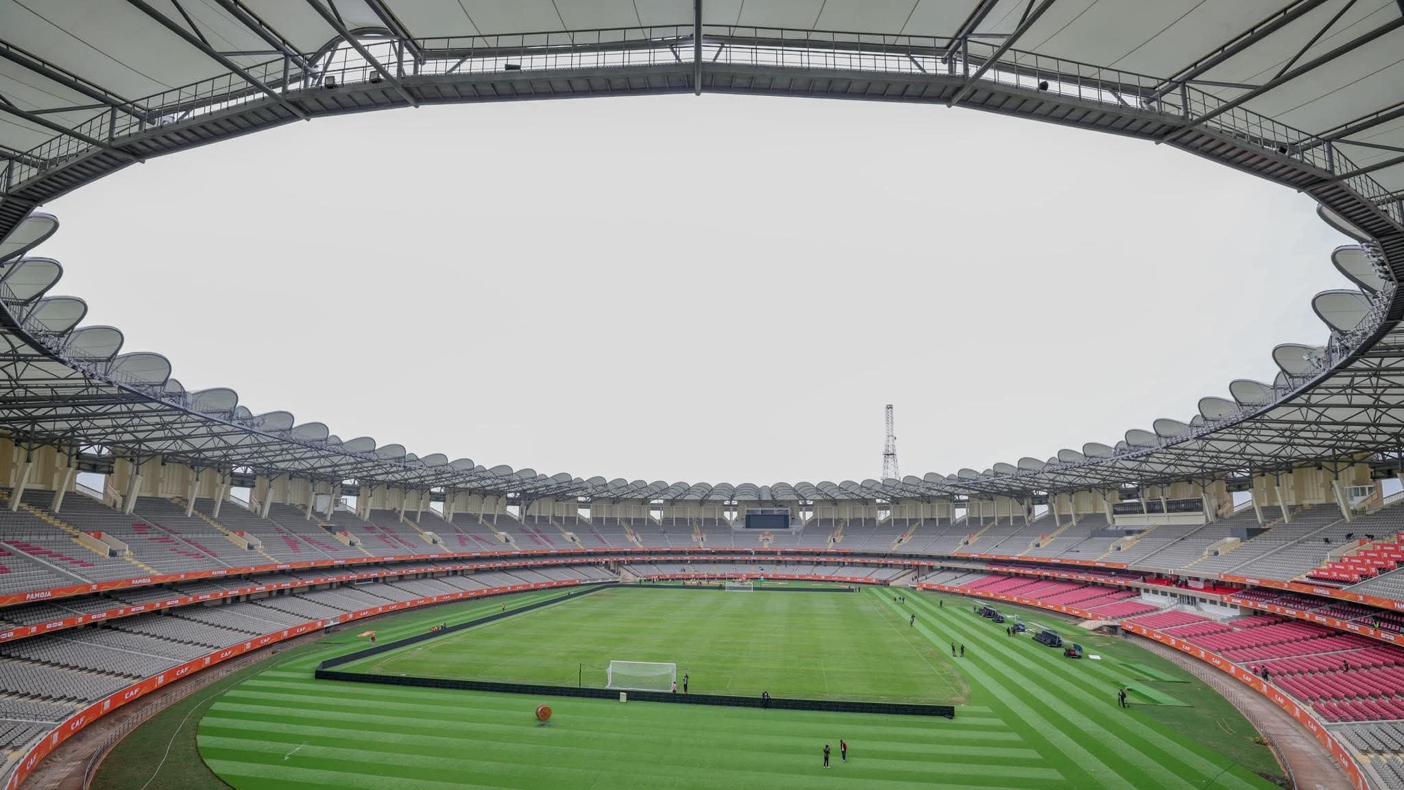 An aerial wide-angle view of the empty interior of the Moi International Sports Centre, Kasarani, showing the green pitch and the red and white seating tiers under a white canopy roof.