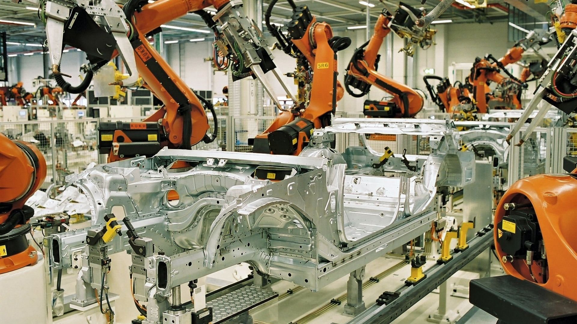 Wide shot of an automated factory floor showing multiple orange robotic arms positioned along an advanced industrial assembly line.