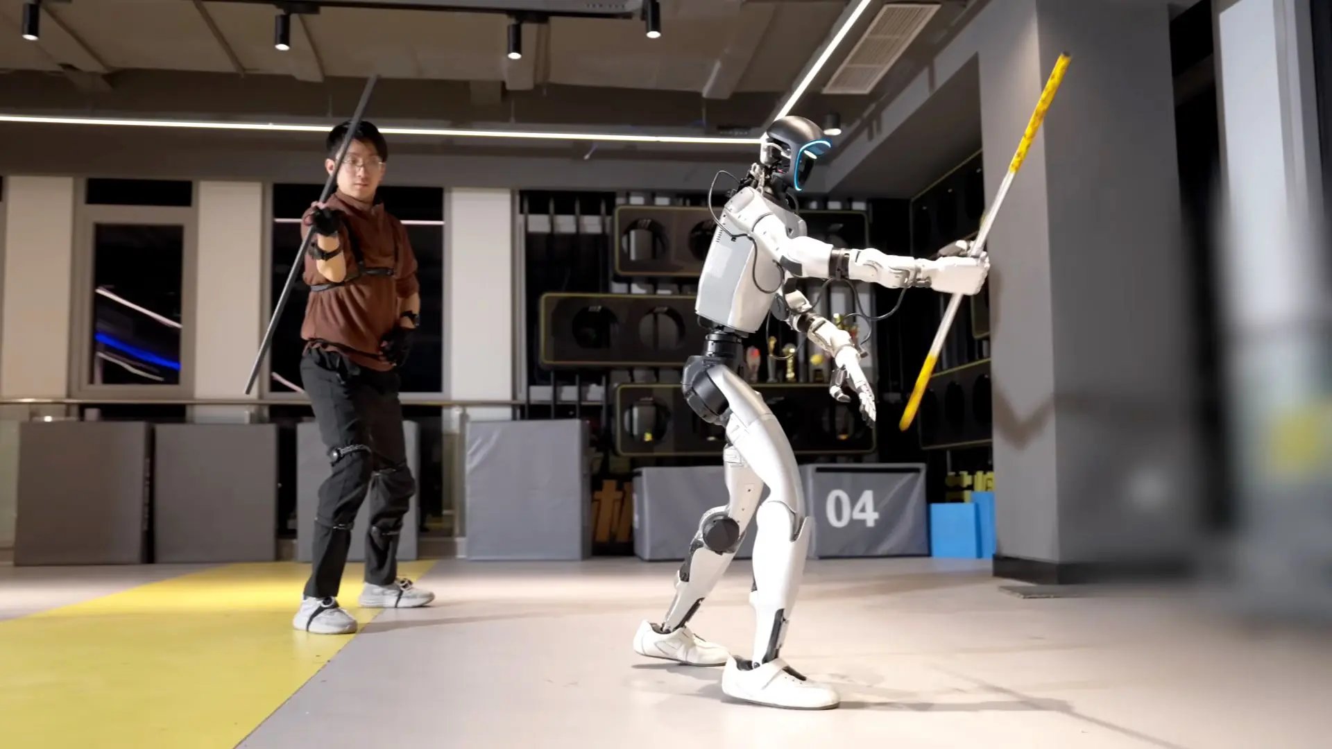 A person wearing a motion-tracking suit poses with a sword opposite the Fuxi humanoid robot holding a training stick inside a testing facility.
