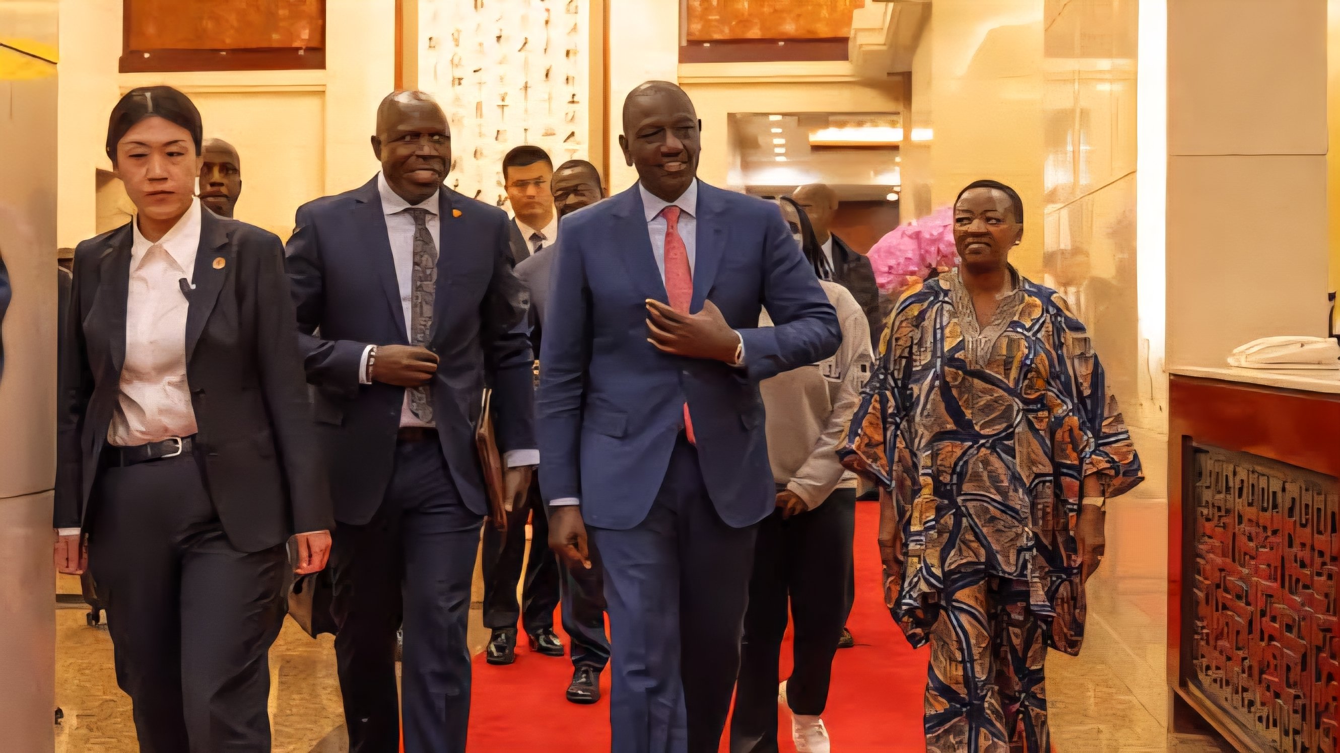 President William Ruto, wearing a blue suit and red tie, walks down a red carpet corridor alongside an entourage of Kenyan and Chinese delegates.