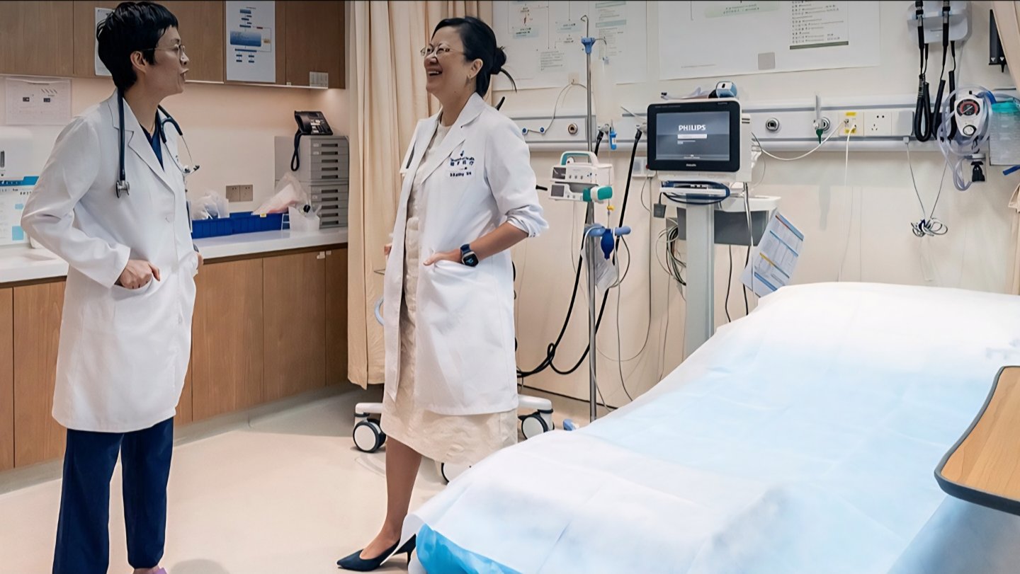Two female medical doctors in white lab coats talking next to an empty hospital bed inside a well-equipped clinical ward in China.