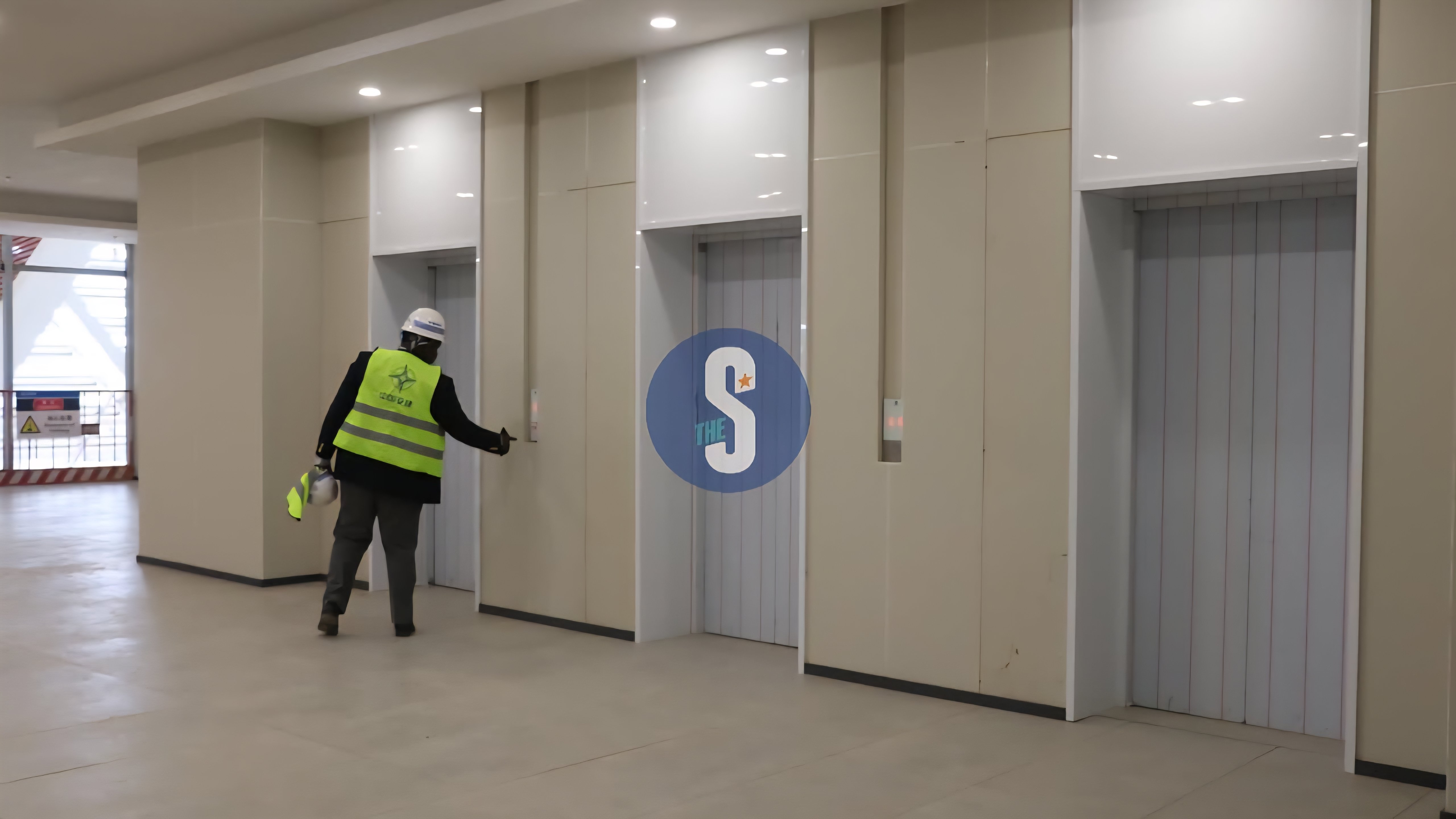 A worker wearing a high-visibility vest and a safety helmet stands in a modern elevator lobby within the Talanta Sports City stadium, pointing at the controls.