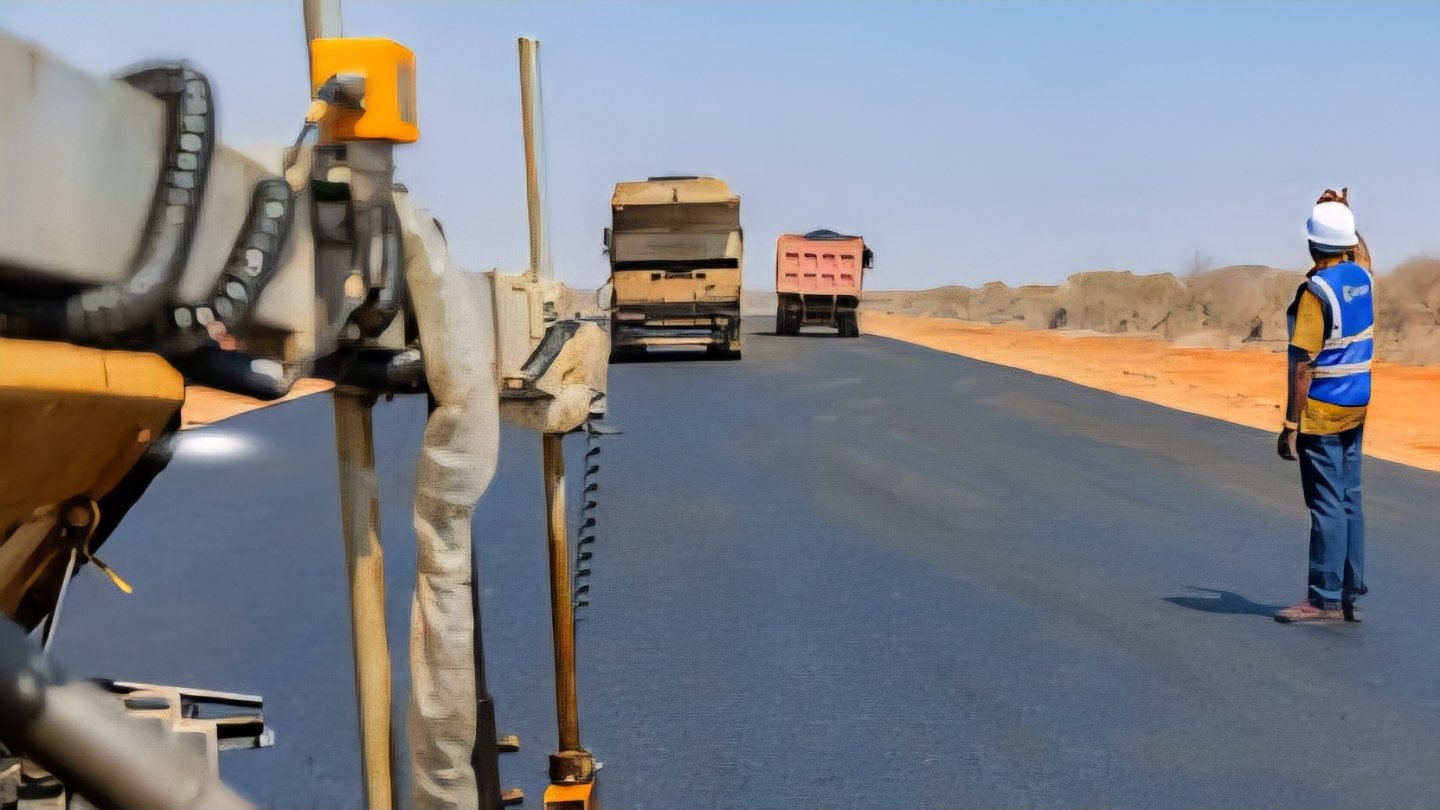 An asphalt paver laying a new road surface on the Mandera-Isiolo road project in Kenya with construction workers and trucks in the background.