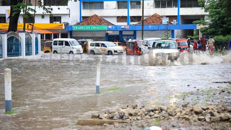 A flooded street in Mombasa during a heavy downpour with people wading through knee-deep water next to a motorcycle and commercial buildings.