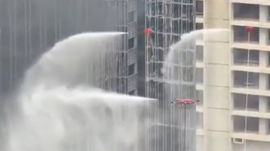Black smoke and flames rise along the exterior glass wall of a tall skyscraper in Shenzhen as emergency drones operate nearby during a high-rise fire test.