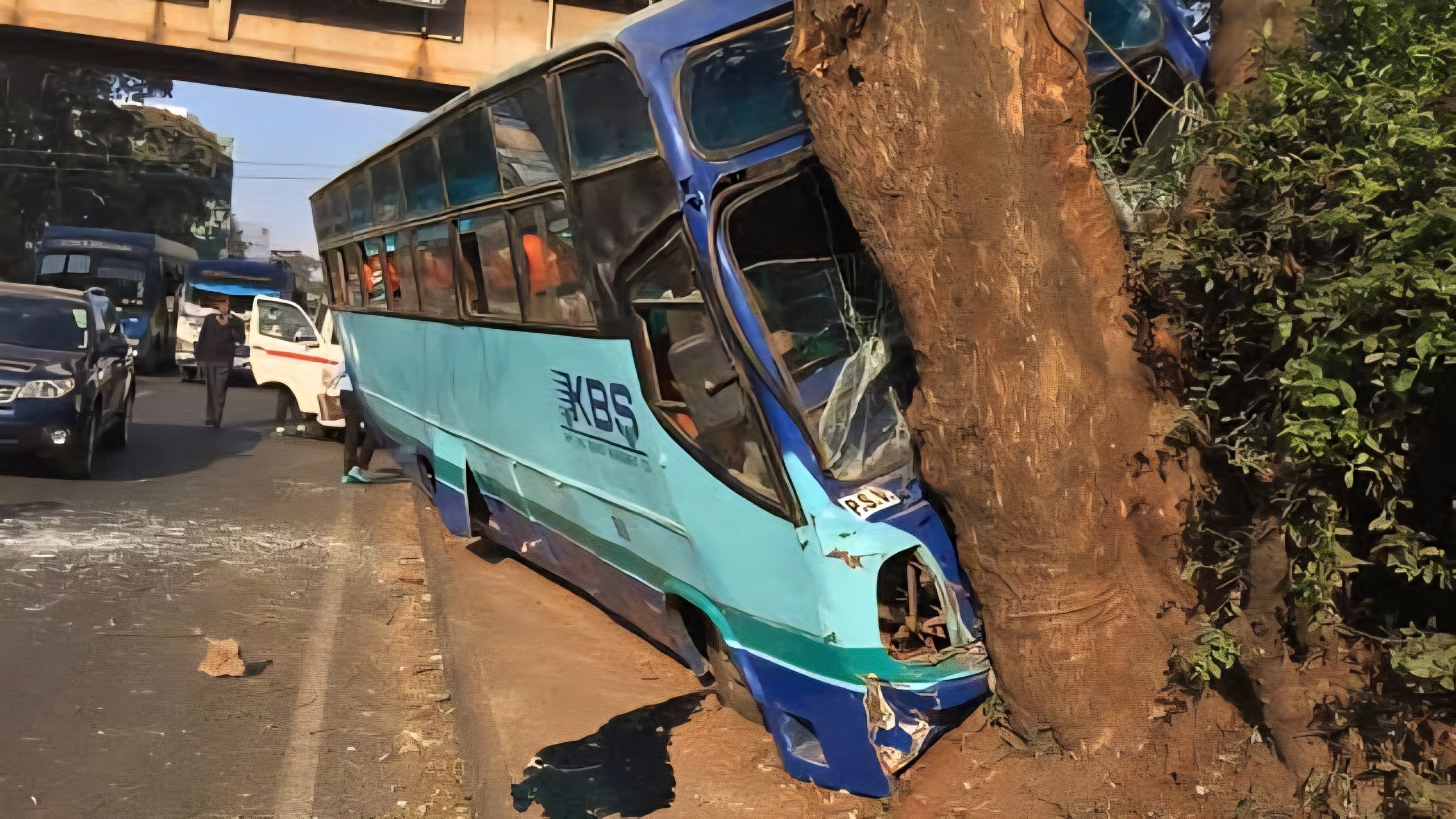The mangled front end of a blue and green KBS bus after colliding with the rear of a stationary commercial lorry on a paved city road.