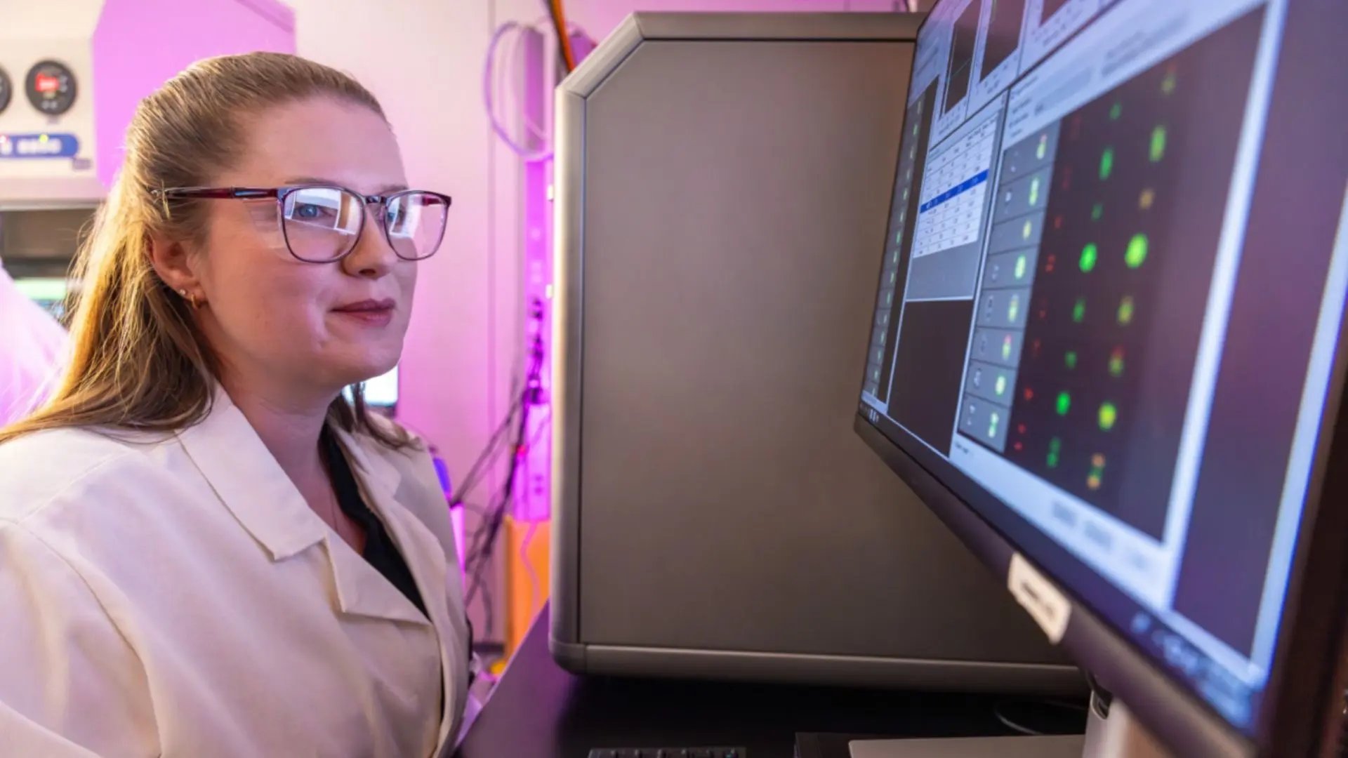 A laboratory researcher wearing protective eyewear operates a desktop computer monitor inside a pharmaceutical facility.
