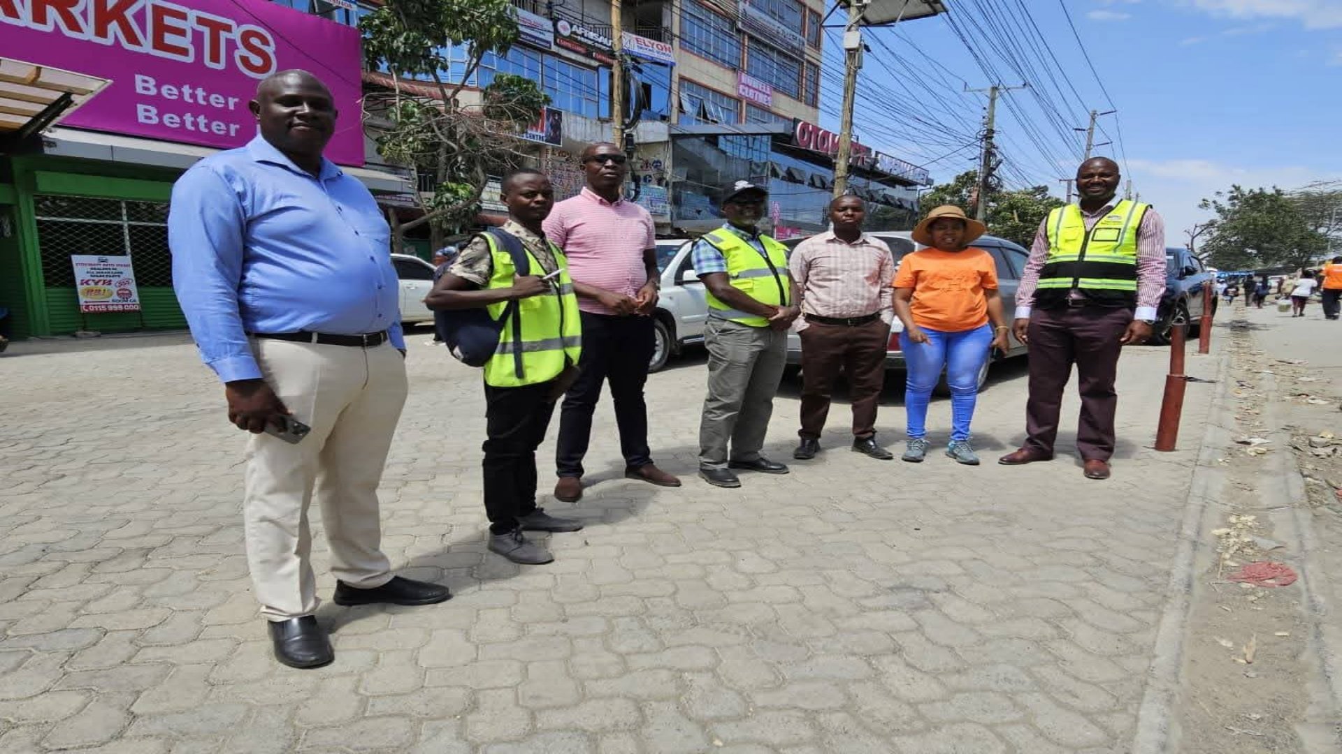 A group of engineering and government officials in professional attire standing outdoors near a paved road surface for a stakeholder meeting.