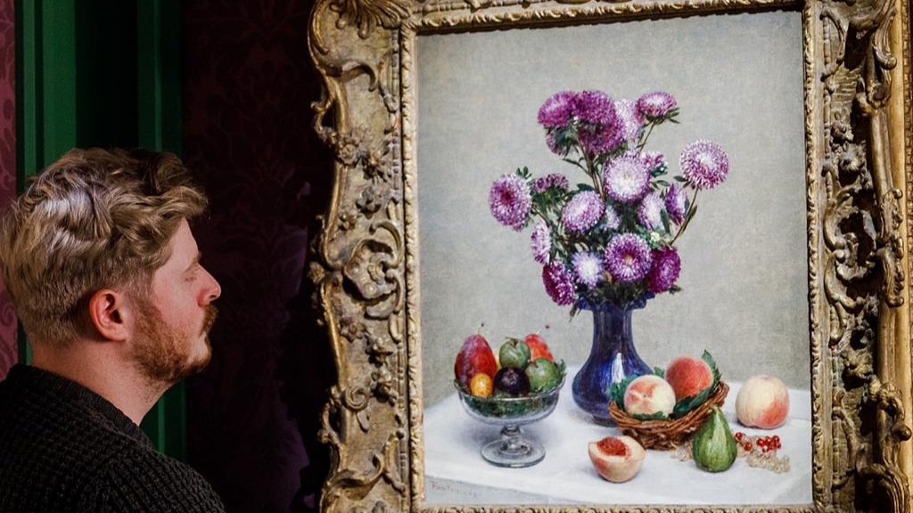 A person views a framed floral oil painting and fruit still-life artwork displayed inside an art gallery or auction space.