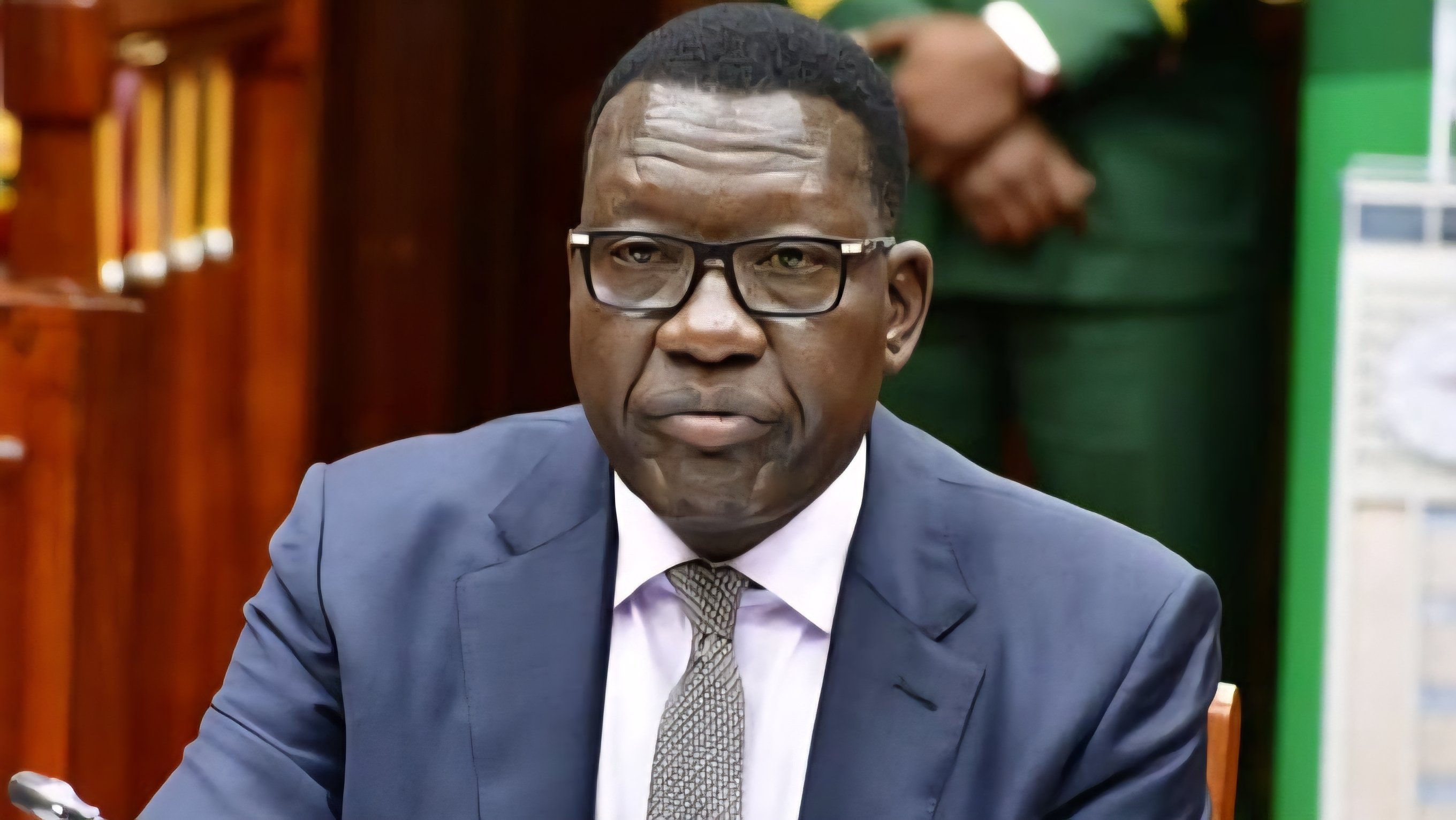 A formal medium shot of a male official in a checked blazer and blue tie, seated in an office chair with a Kenyan flag in the background.