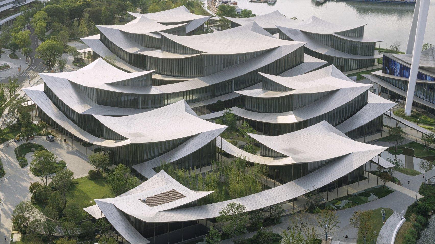 Aerial view of the Suzhou Museum of Contemporary Art on the banks of Jinji Lake in China, showing twelve interconnected pavilions beneath a continuous rippled stainless steel roof surrounded by landscaped gardens.