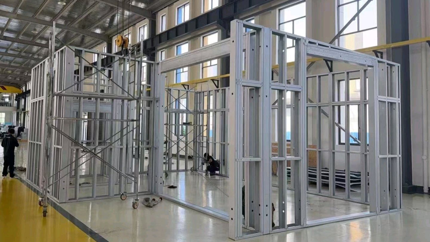 A factory workshop floor showing a partially assembled light gauge steel wall frame structure with scaffolding nearby.