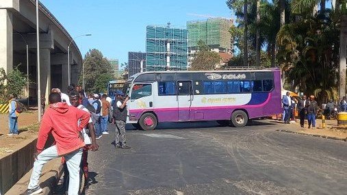 Chaotic Nairobi traffic under a bridge with smoke, gridlocked colorful matatus, police, boda bodas, and crowds.