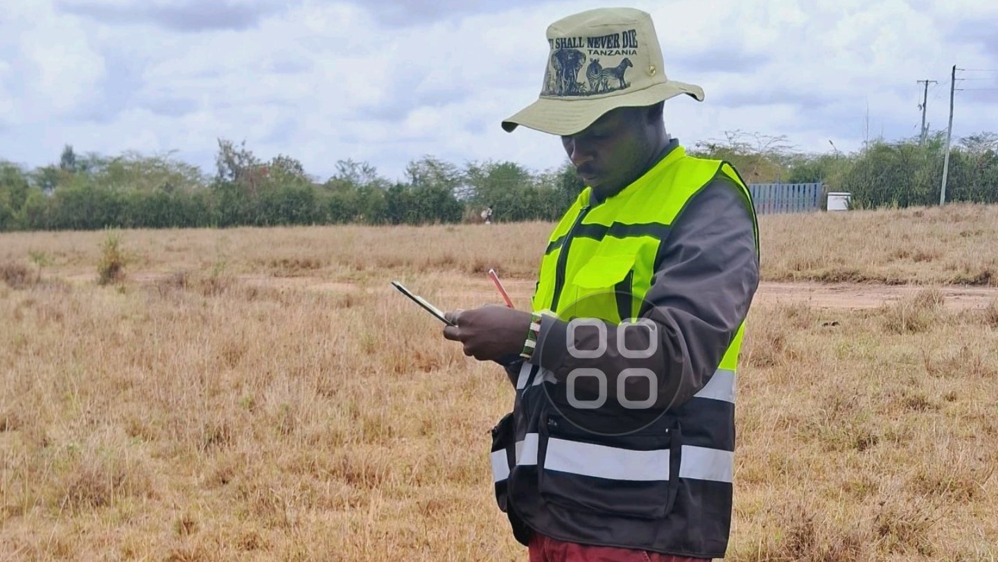 A professional surveyor standing in an open grassy field, wearing a high-visibility safety vest and branded hat while documenting field measurements on a mobile device, as shown in file 259794.png.