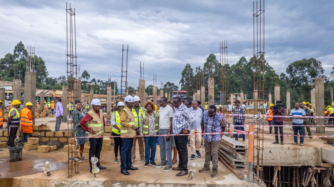 Wide shot of President William Ruto wearing a white hard hat and yellow reflective vest, standing with officials at the Nyakoe Modern Market construction site in Kisii.