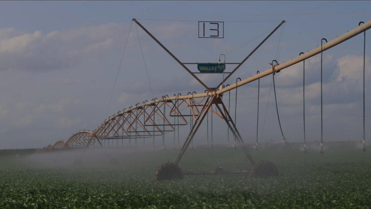 A large mechanical pivot irrigation system spraying water over a green agricultural field under a cloudy sky.