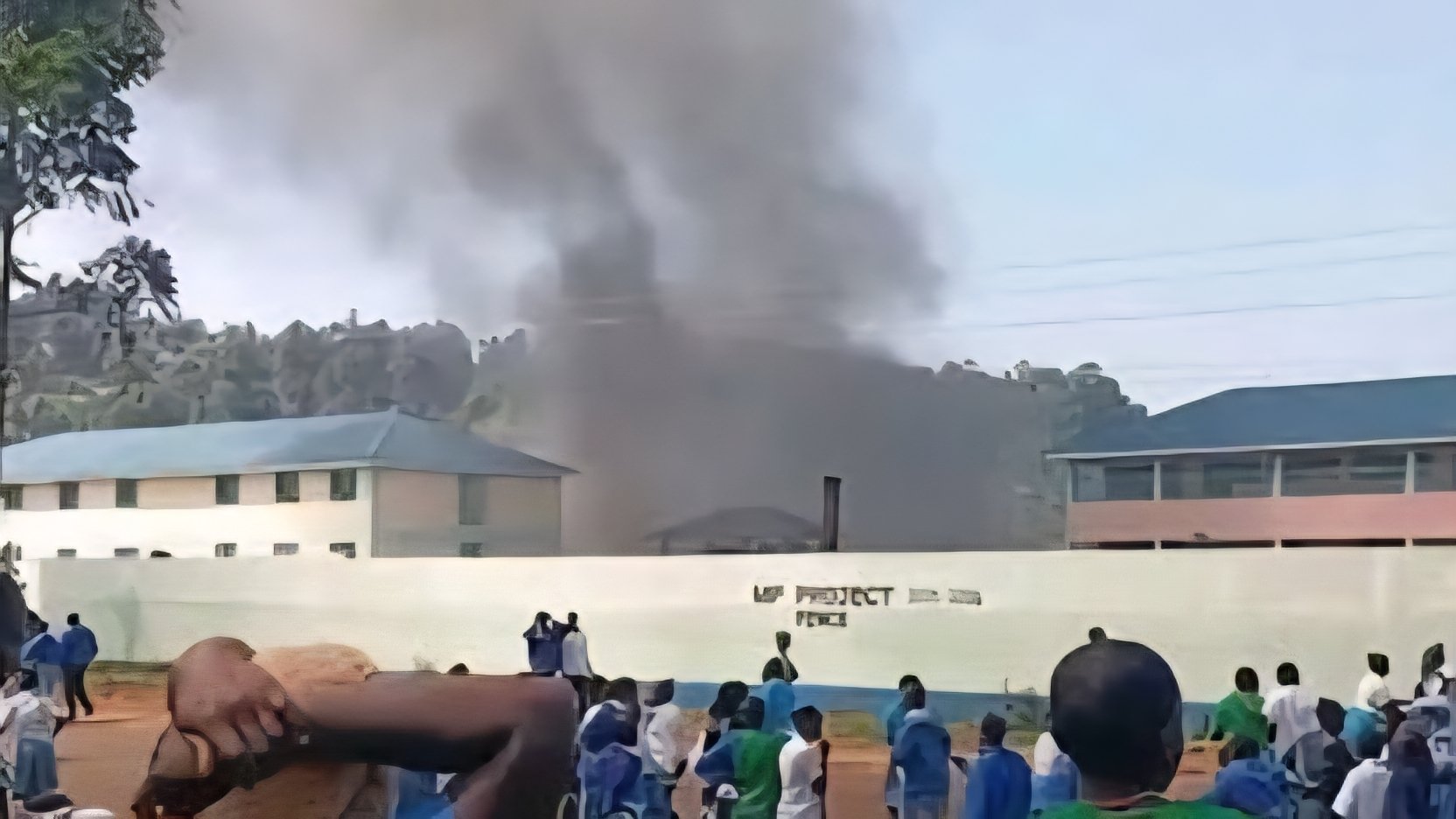 A thick plume of dark smoke billowing into the sky from a burning school dormitory building behind a concrete perimeter wall.