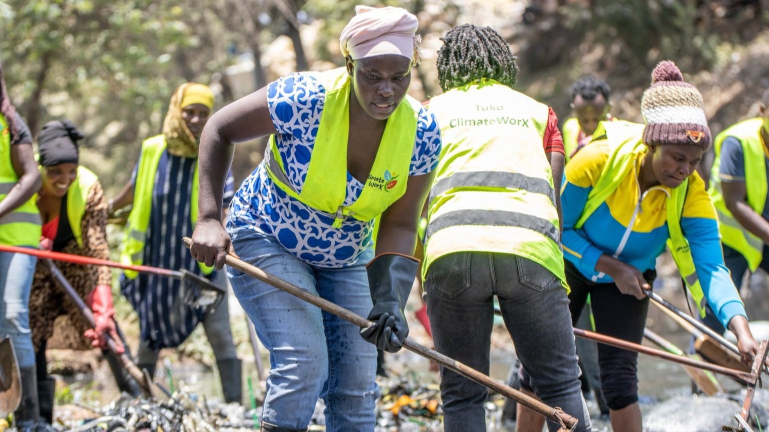 Nairobi City County sanitation workers clearing debris from an open drainage system along a city street.