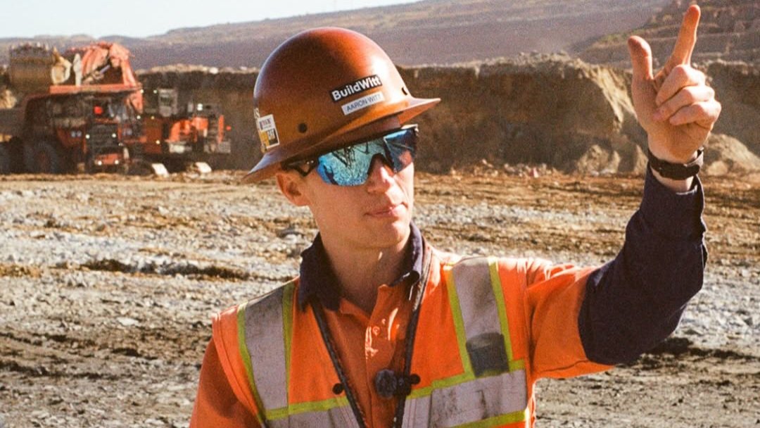 Aaron Witt wearing a helmet and safety equipment gestures toward heavy earthmoving machinery at an open excavation site.