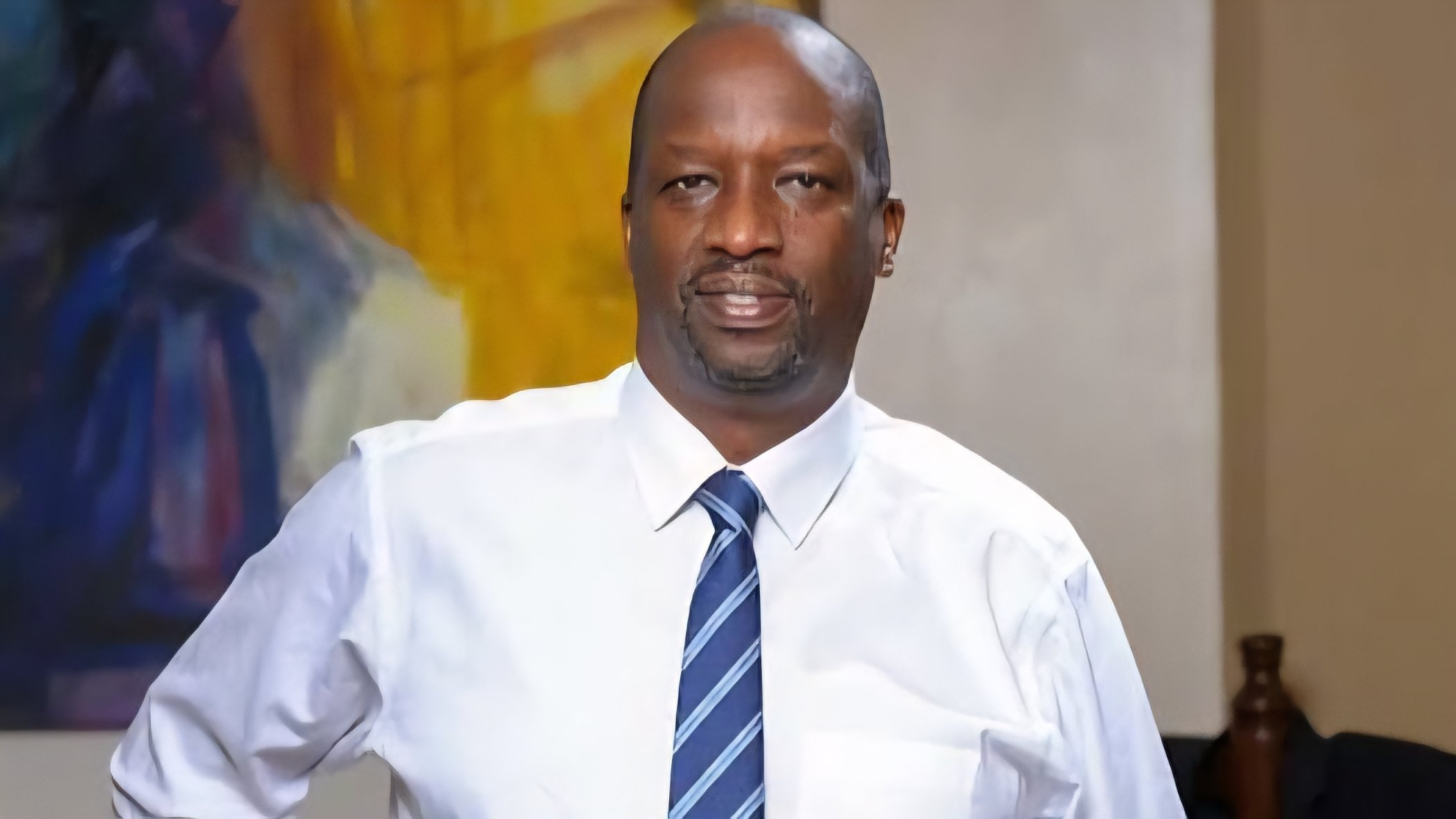 A formal portrait of Kiprono Kittony wearing a white shirt and blue striped tie against a blurred background.