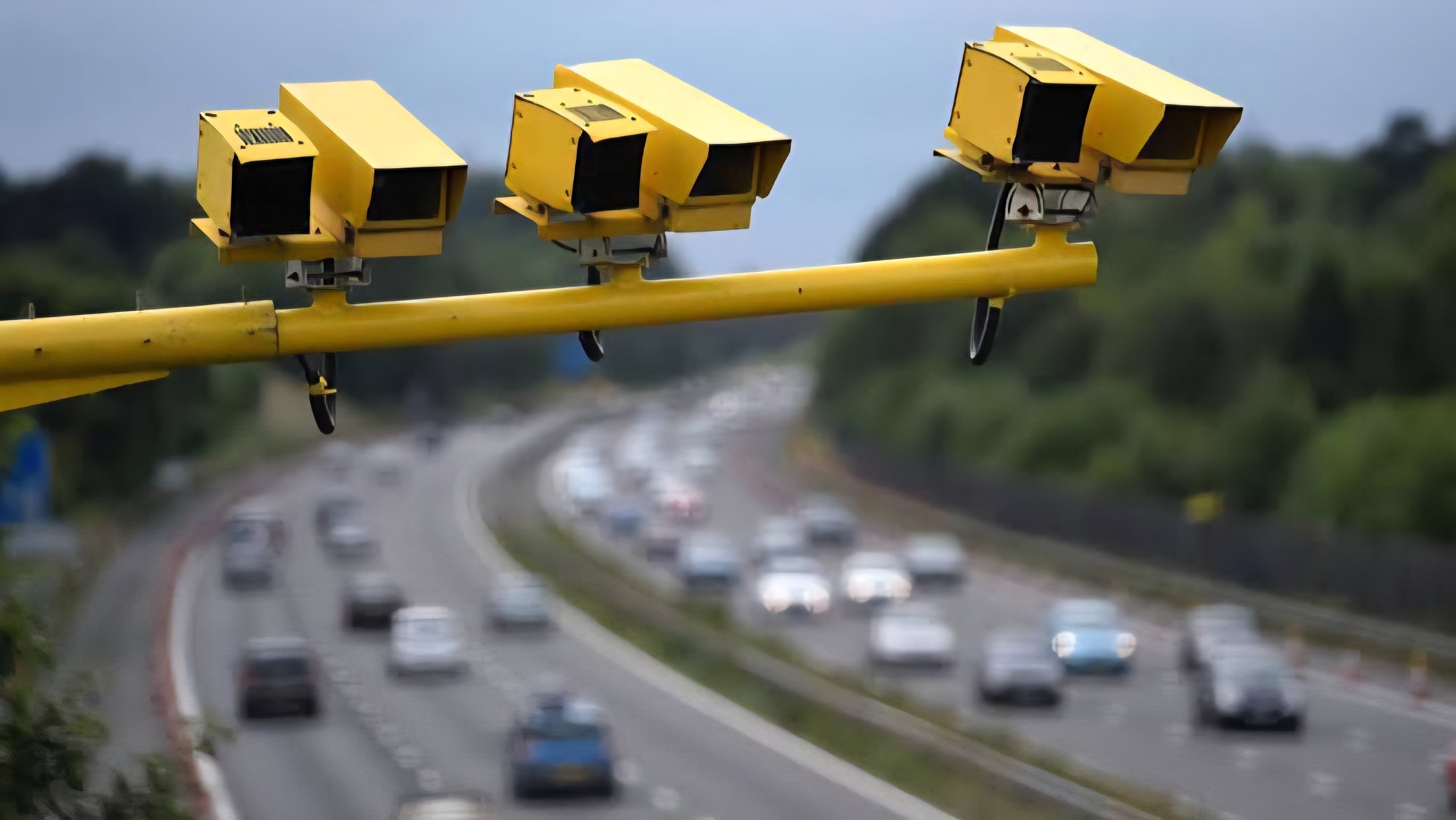 Three bright yellow traffic enforcement cameras mounted on an overhead yellow metal gantry overlooking a busy multi-lane highway with blurred moving vehicles.