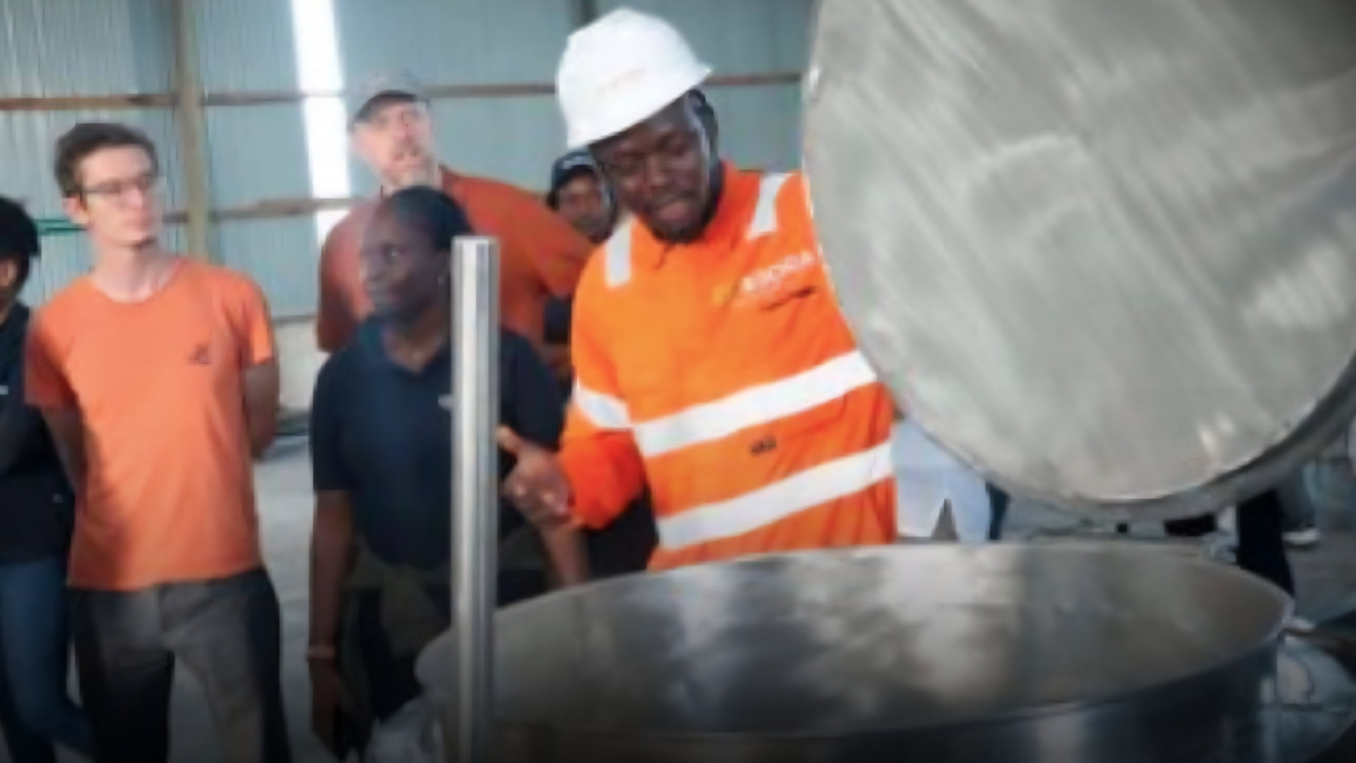 A Kenyan chef in an orange high-visibility jacket demonstrates a large industrial electric pressure cooker to onlookers at a school kitchen facility.
