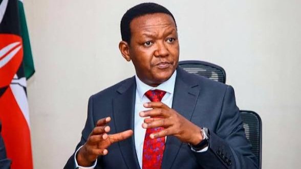 A man in a navy blue suit and red tie speaks at a wooden podium during a formal government briefing or press conference.