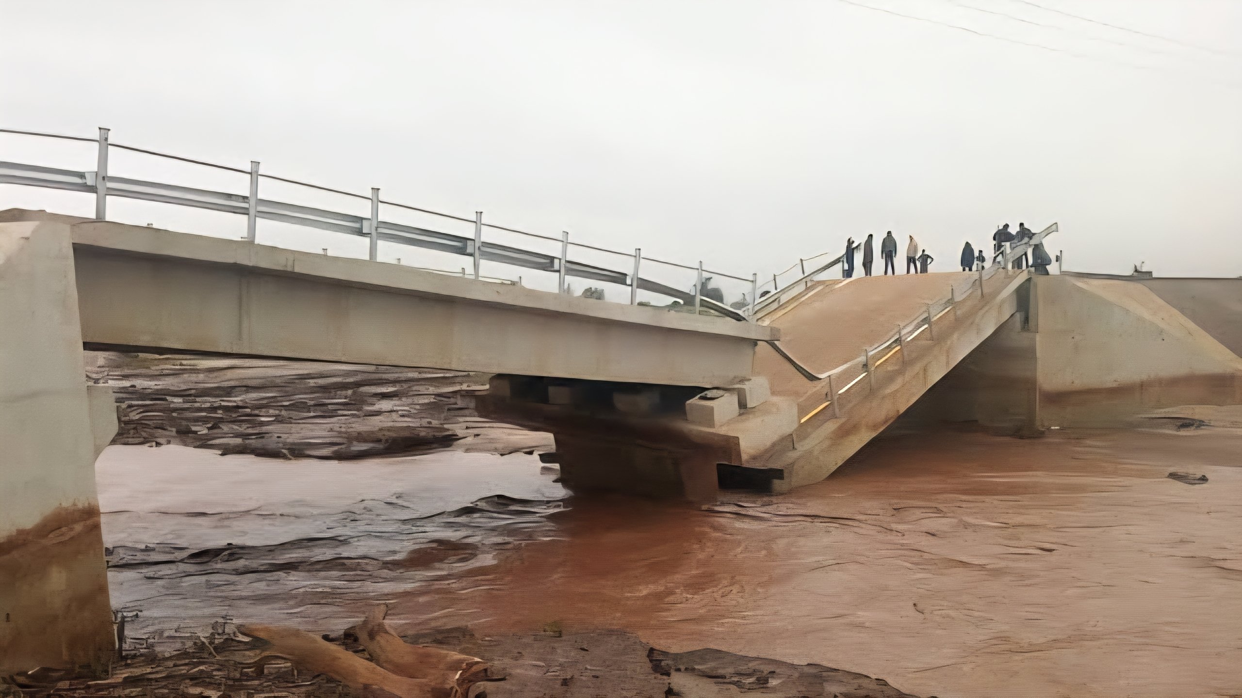 A flooded road in Kenya showing a collapsed bridge section and brown, stagnant water filling a large excavation site near a construction area.