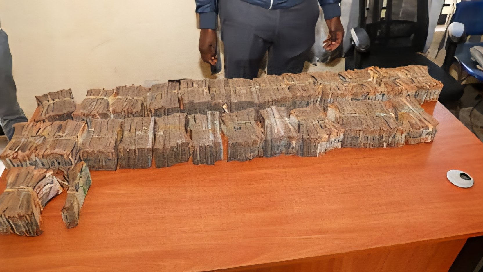 A high-angle medium shot shows several long rows of bundled Kenyan shilling bank notes stacked neatly across a wooden office desk during a corruption investigation inventory.