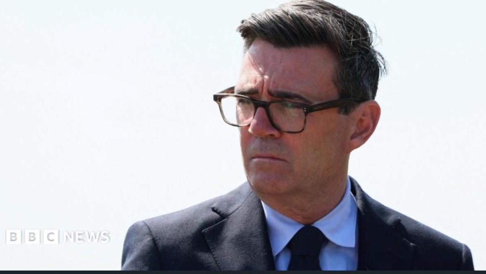 Prime Minister Andy Burnham delivering an official address outdoors in a dark suit and glasses.