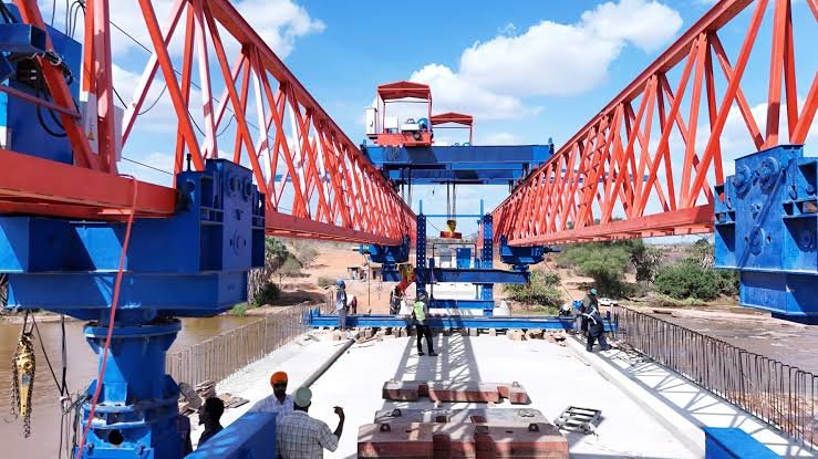 An aerial view of the nearly completed concrete bridge spanning the Galana River, showing construction equipment and the expansive irrigation fields of the Galana Kulalu scheme in the background.