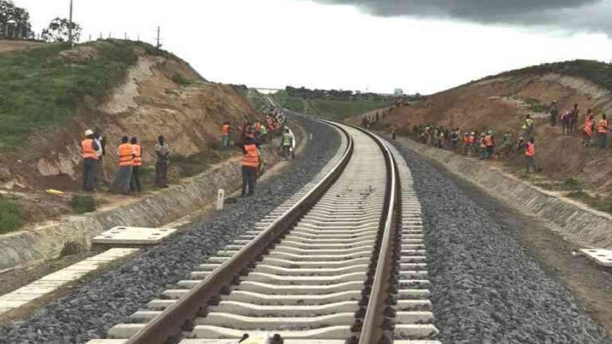 A long-distance view of a Standard Gauge Railway track cutting through a rural Kenyan landscape with a cargo train in the distance.