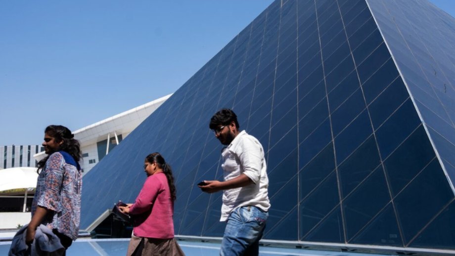 Pedestrians walking past a modern building with a sharp, geometric blue glass facade, reflecting the sky.