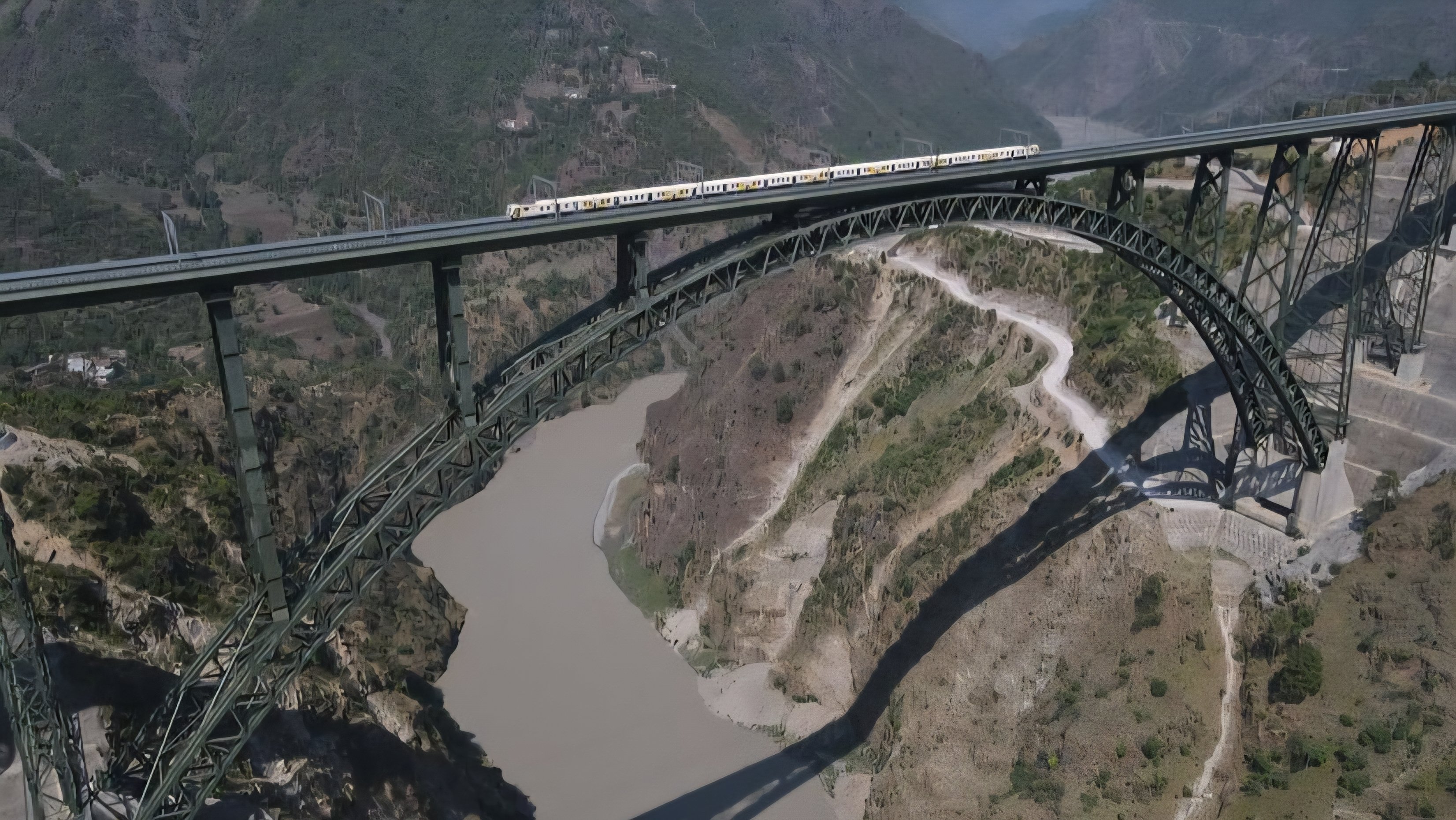 High-angle view of a passenger train traversing the high-elevation steel arch Chenab Railway Bridge over a river gorge.