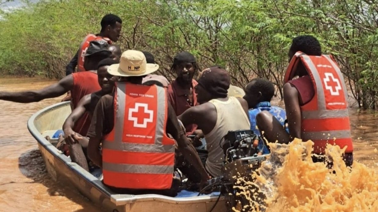 A red inflatable rescue boat carrying several people, including a Red Cross volunteer, through brown floodwaters in an area with submerged trees.