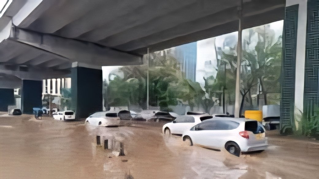 A wide brown river of floodwater flowing through a damaged urban road in Nairobi, with a collapsed bridge pillar in the center and buildings in the background.