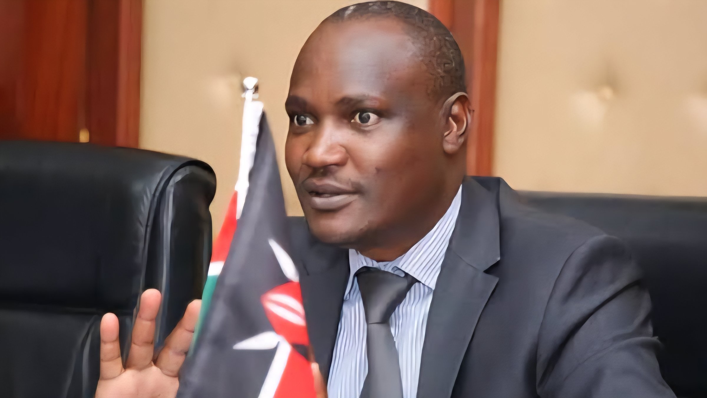 Close-up photo of a Kenyan government official in a suit, speaking at a podium with water bottles and a microphone visible during a budget presentation.