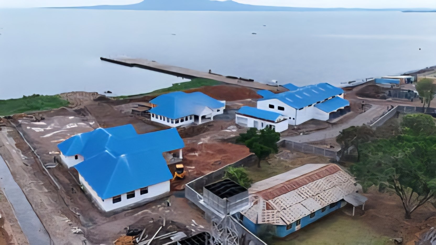 An aerial view of the Homa Bay Pier construction site on Lake Victoria, showing new white buildings with blue roofs, a long concrete jetty extending into the water, and organized warehouse structures.